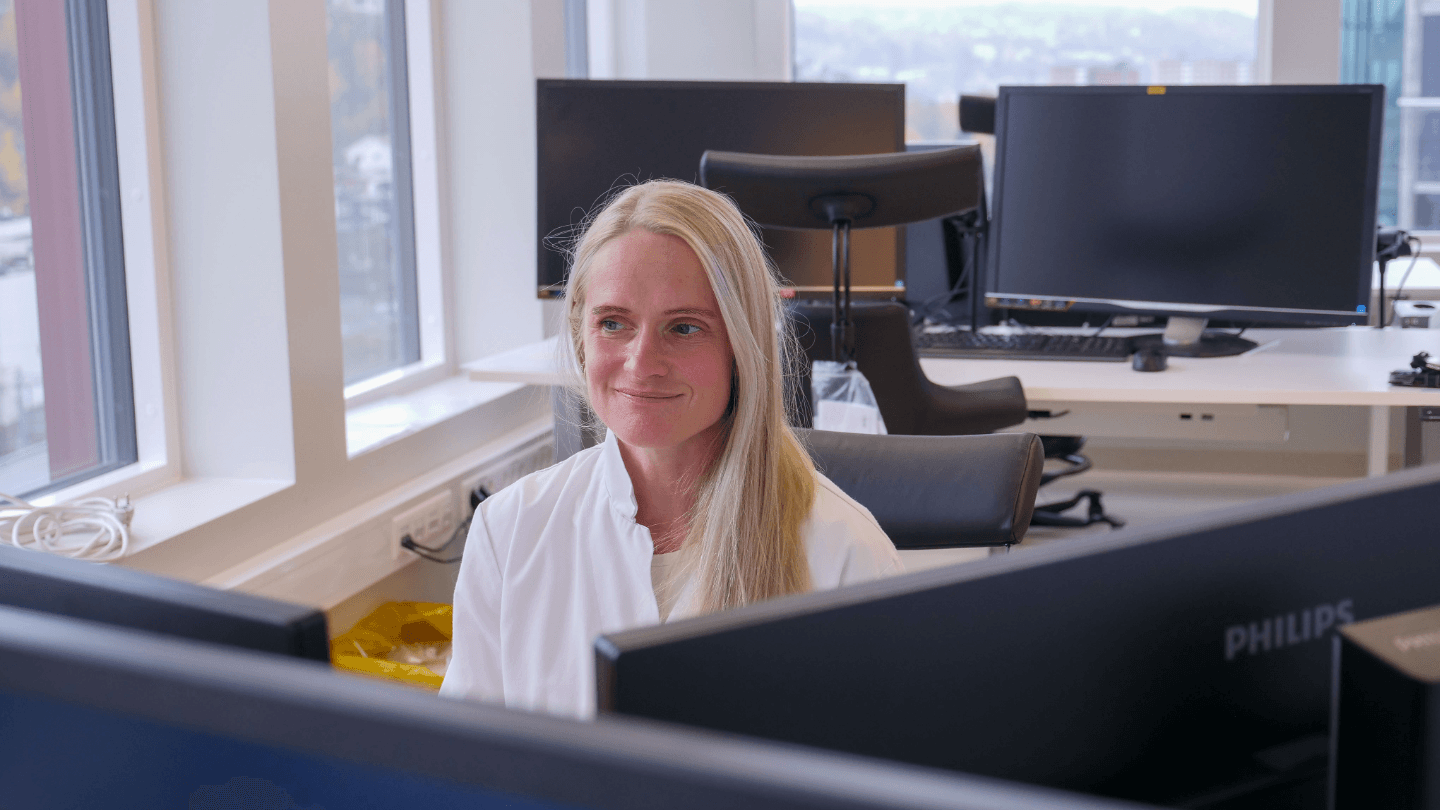 Kvinne sitter foran to dataskjermer i et kontorlandskap.