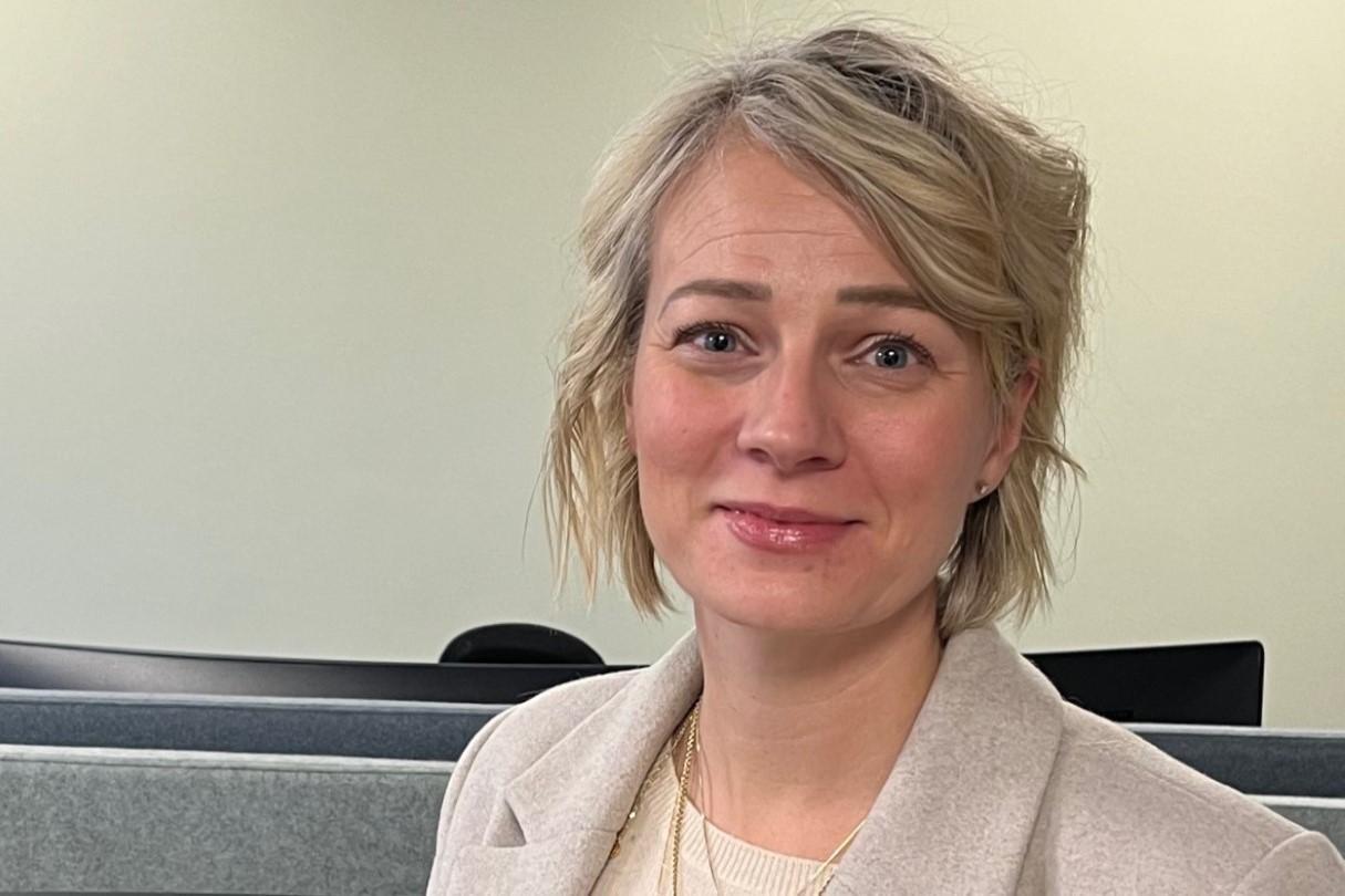 Lene står foran en kontorplass. På skrivebordet er det en bred skjerm. Lene har kort, lyst hår. Hun har på seg en beige dressjakke.