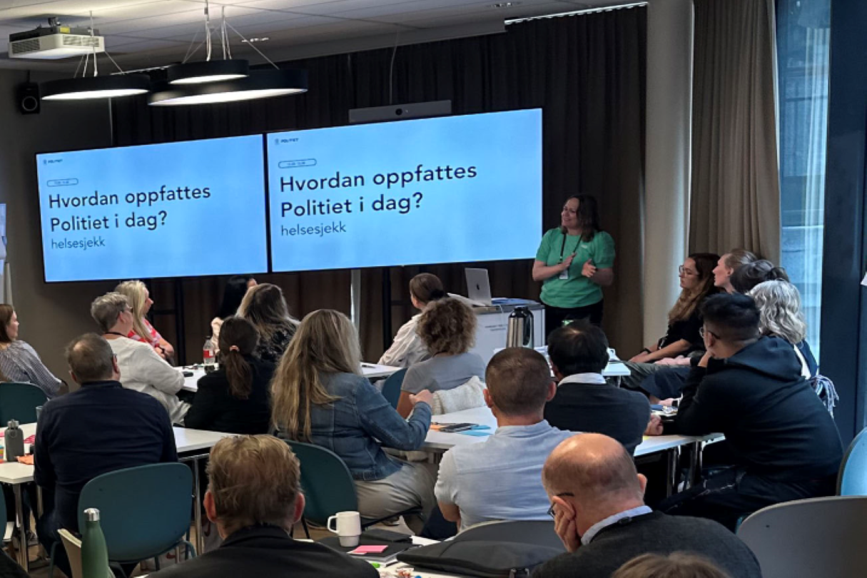 Ansatte som sitter i et rom under workshop. De sitter og ser på en skjerm og hører på foredragsholder.