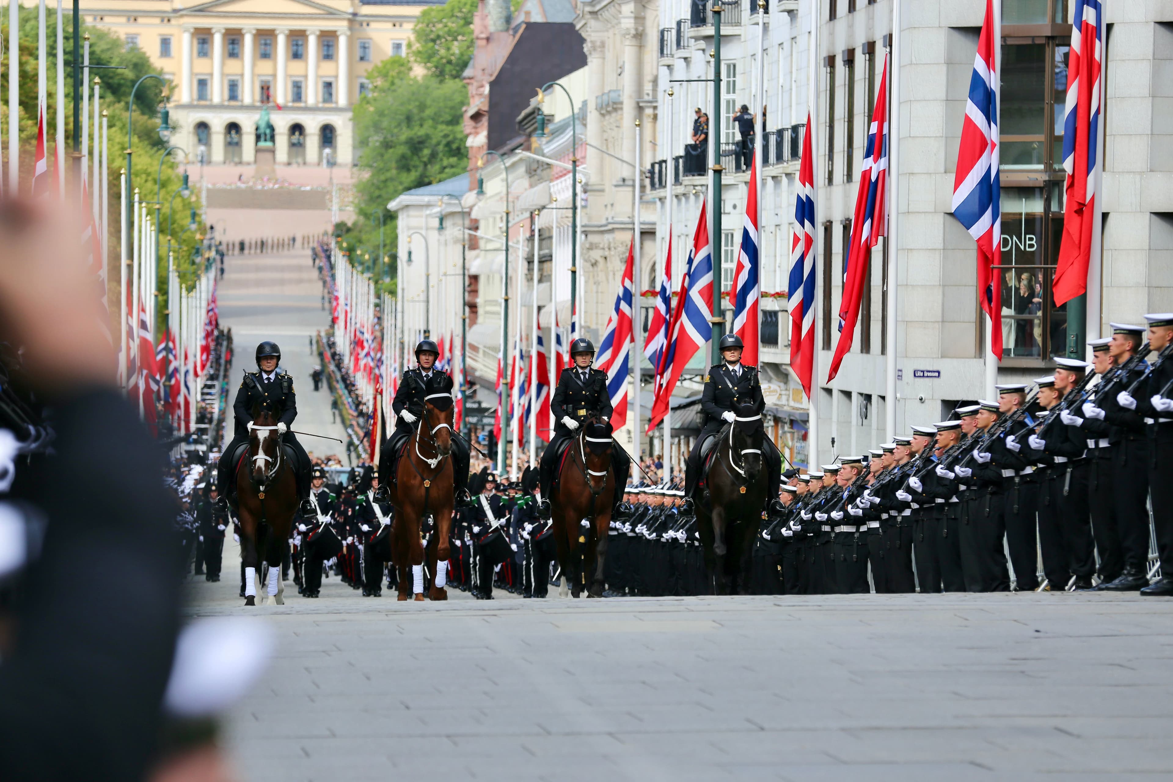 4 politihester rir opp Karl Johan i Oslo. Publikum og soldater fra Forsvaret står langs siden. Flagg på halv stang på hver side.